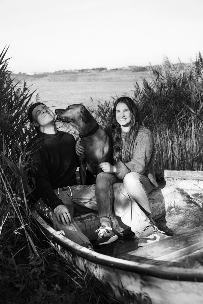 Black-and-white dog photography in Vejers: couple in wooden rowboat, dog licks partner's cheek, reeds, smiling woman.