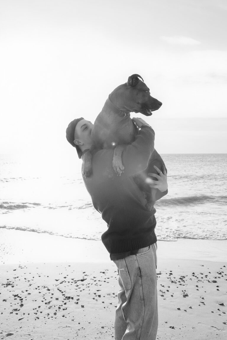 Hundefotografie in Henne: Hundebesitzer im Pullover trägt den Hund am Strand im Gegenlicht.