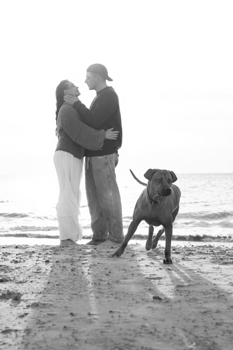 Hundefotografi i Blåvand: par omfavner på stranden, hund i forgrunden ved Vesterhavet