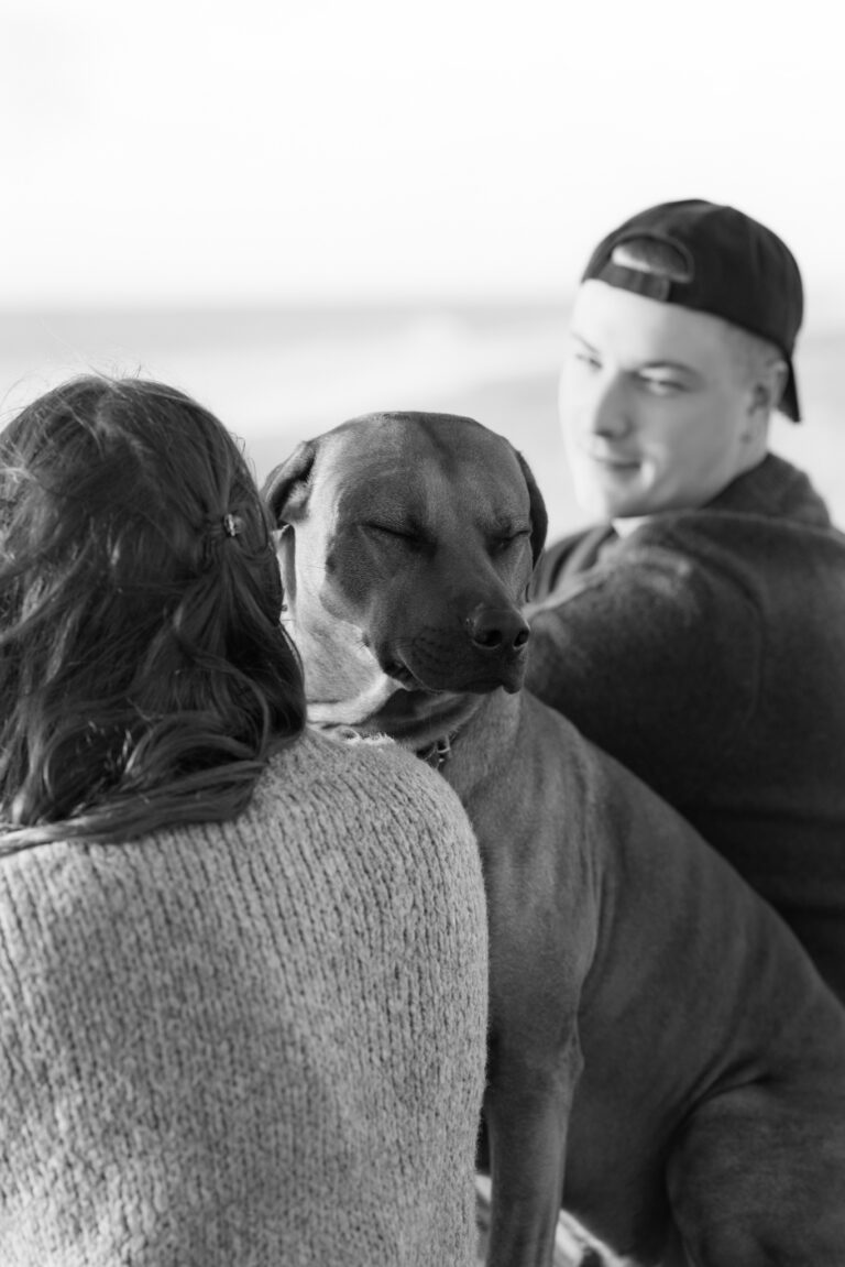 Sort-hvidt hundefotografi fra Henne: hund med lukkede øjne læner sig mod en kvinde i strik, mens en mand i kasket ser på.
