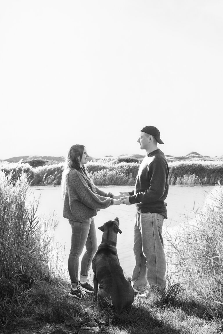 Hundefotografie in Hvide Sande: Paar hält Hände, dunkler Hund sitzt zwischen ihnen, Schilf und Wasser.