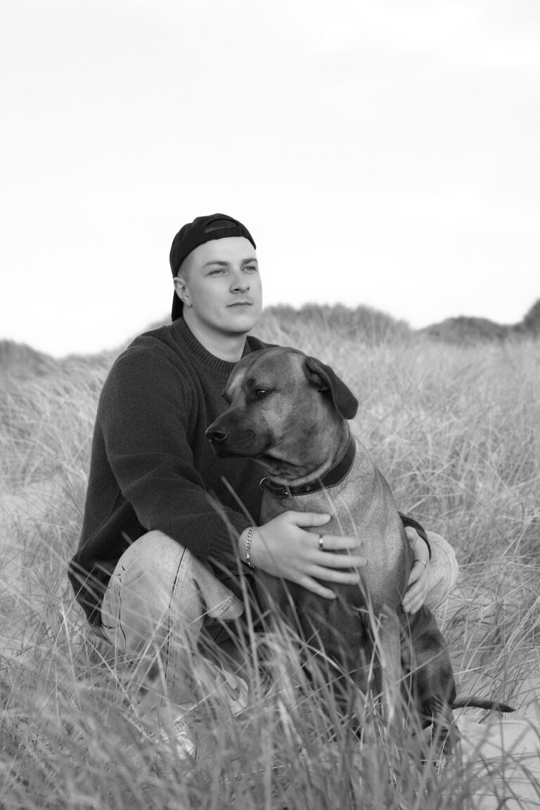 Black-and-white dog photography in Henne: dog owner kneeling in dune grass, embracing a large short-haired dog.