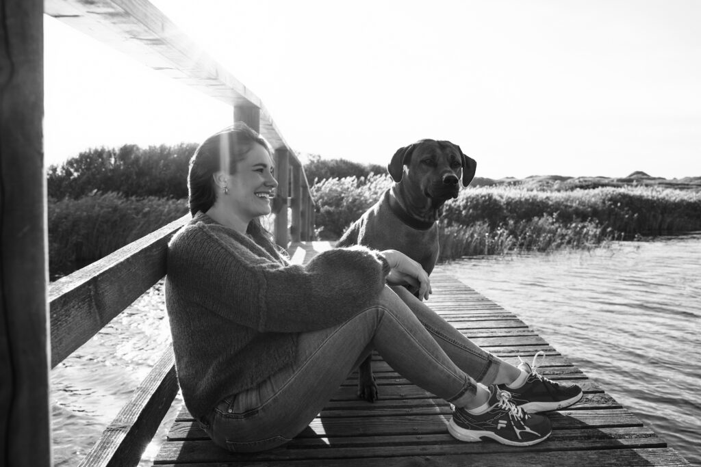 Schwarz-Weiß-Hundefotografie in Hvide Sande: Frau im Strickpullover mit Hund auf Holzsteg.
