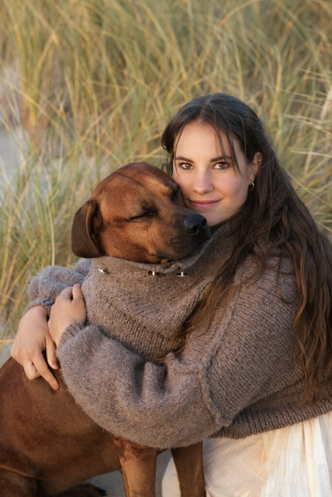 Hundeejer med langt hår i brun sweater krammer en brun hund i klitgræs ved Blåvand under hundefotografering.
