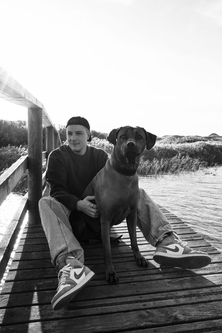 Sort-hvidt hundefotografi fra Vejers: hund og hundeejer sidder på en træbro over vandet.