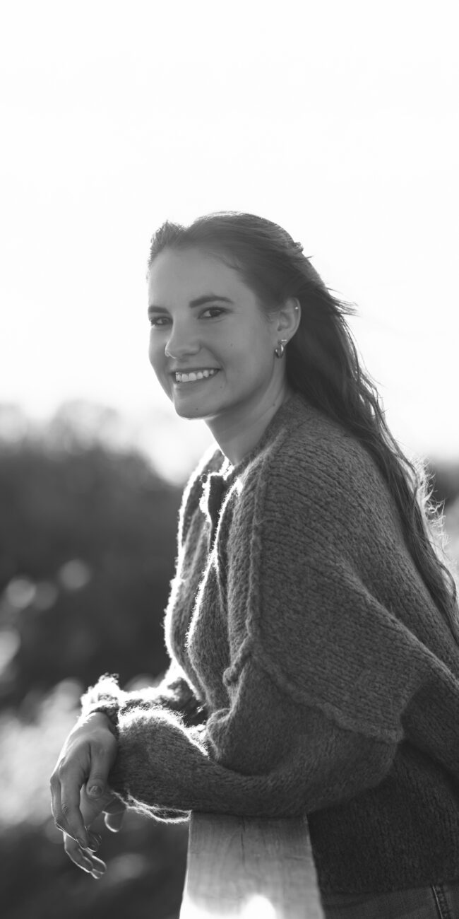 Black and white dog photography portrait in Henne: smiling woman in knitted sweater leaning on fence, backlit.