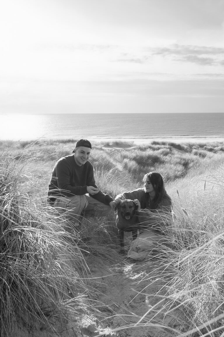 Black-and-white dog photography in Henne: couple with dog in dune grass, North Sea horizon behind.